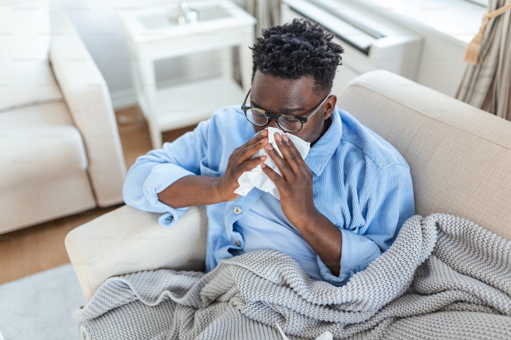 Ill African young man covered with blanket blowing running nose got fever caught cold sneezing in tissue sit on sofa, sick allergic black man having allergy symptoms coughing at home, Corona 19