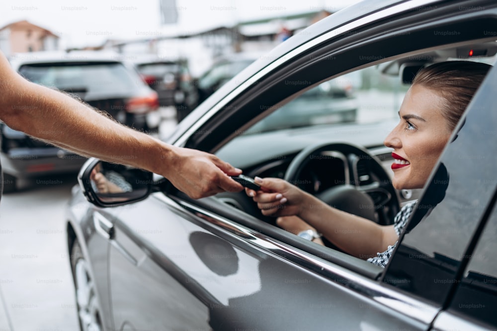 Acquirente femmina soddisfatto e sorridente seduto nella sua nuova auto. Sorride, guarda il venditore attraverso la finestra aperta mentre gli prende le chiavi dell'auto.