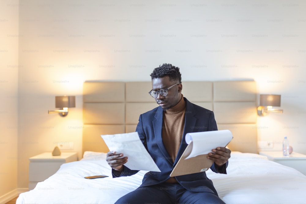 ホテルのベッドに座って仕事をしている真面目なアリカ人ビジネスマンは、ホテルの部屋で出張の費用を分析します。