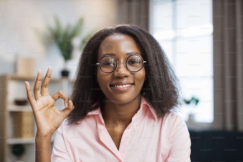 Smiling african american woman in eyeglasses and pink shirt sitting at desk with books and notes and making okay. Female tutor working at home during distance learning.