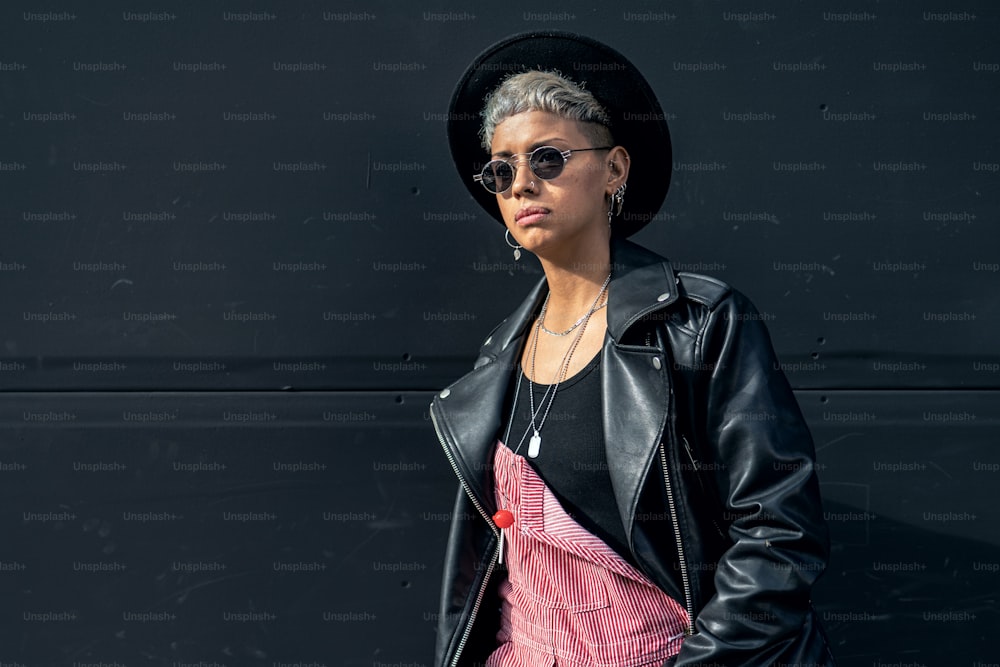 Mujer joven moderna de moda con gafas de sol, sombrero negro y chaqueta, posando en la muralla negra de la ciudad. Mucho espacio de copia.