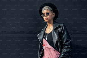 Fashionable modern young woman wearing sunglasses, black hat and jacket, posing on the black city wall. A lot of copy space.