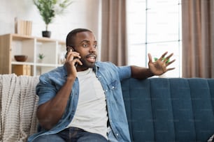 快適なソファで休んでいる間、スマートフォンで話しているカジュアルウェアの��ハンサムなアフリカの男性。家での自由時間にモバイルで会話をしている若い男。