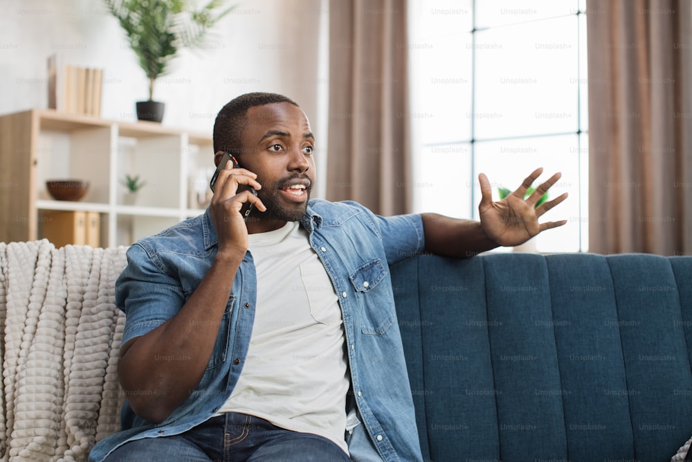 Hübscher afrikanischer Mann in Freizeitkleidung, der auf dem Smartphone spricht, während er sich auf einer bequemen Couch ausruht. Junger Mann, der sich in der Freizeit zu Hause mobil unterhält.