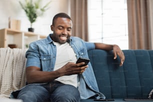 カジュアルウェアを着たアフリカ系アメリカ人の男性が、驚いた表情でモバイル画面を見ています。ソファに座ってモバイルで良いニュースを読んでいる幸せな若い男。