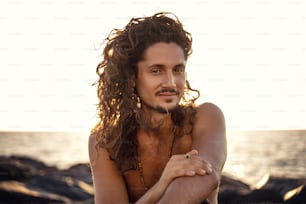 Retrato de un guapo hippie italiano con el pelo largo relajándose en la costa, hora de la puesta del sol. Ojos tranquilos. Hombres mirando a la cámara.