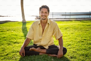 Homem italiano hippie sorridente sentado em lótus, praticando yoga ao ar livre em dia ensolarado. Homens olhando para a câmera com um sorriso bonito. Muito espaço de cópia.