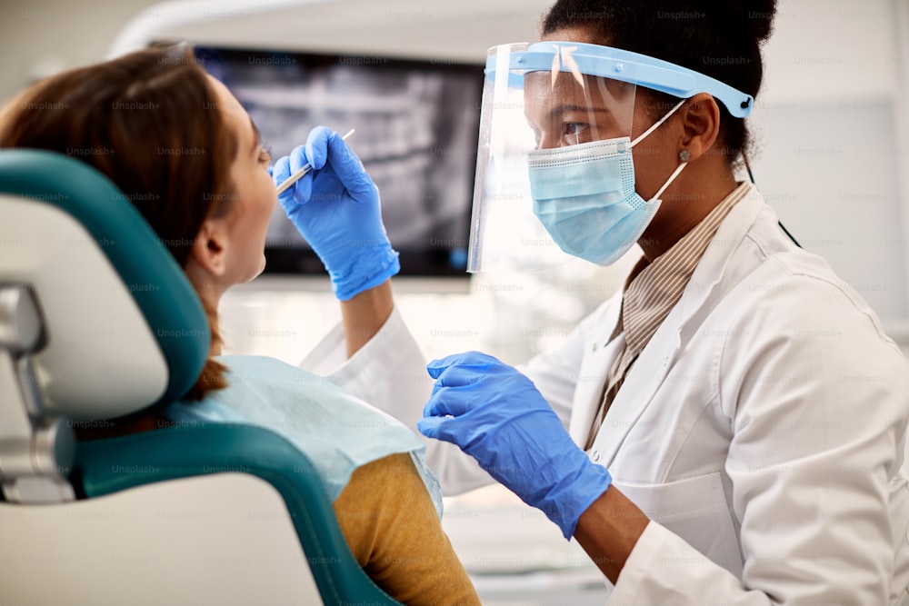 Dentista afro-americano com roupas de trabalho de proteção realizando exame médico em paciente do sexo feminino em clínica odontológica.