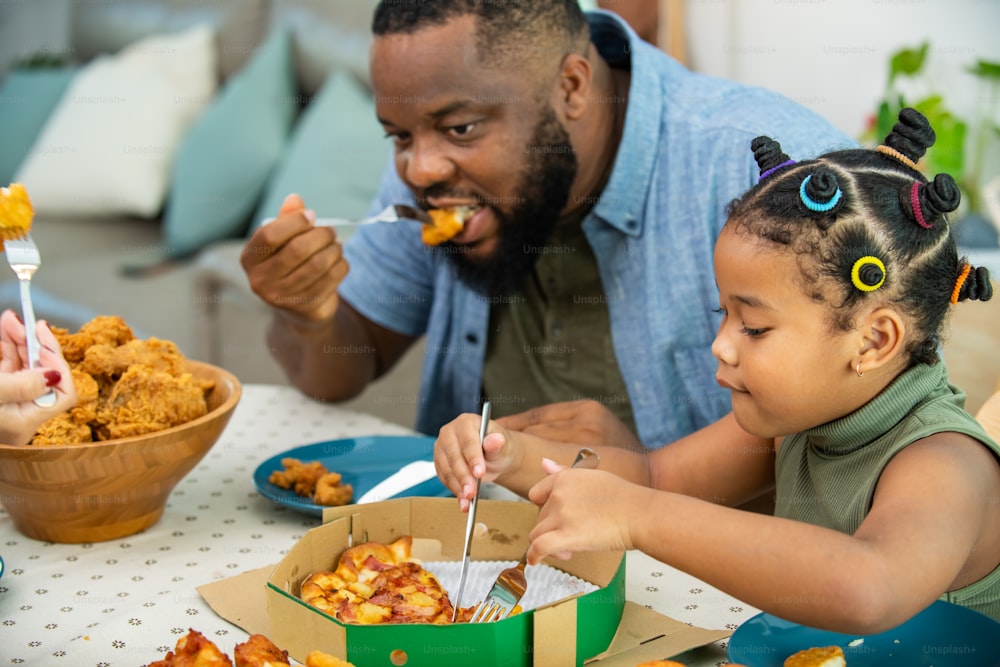 Happy African family parents and two little daughter eating fried chicken and pizza for dinner together. Father and mother and cute child girl kid enjoy eating and sharing a meal together at home
