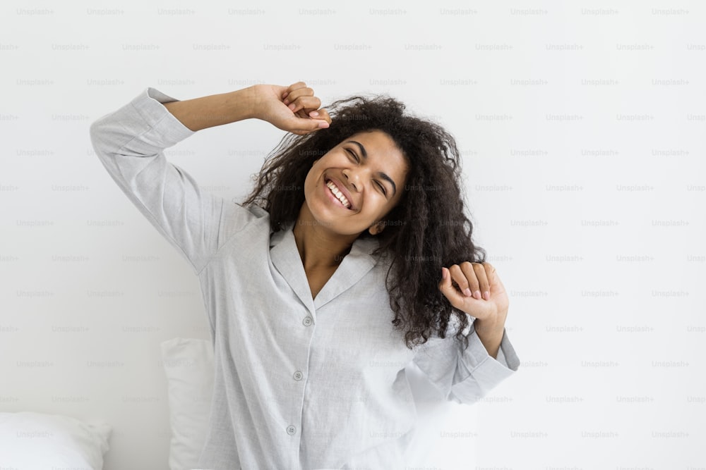 Good morning concept. Happy afro american woman waking up at home, sitting on bed in pajamas, stretching body, raised hands up and smiling wide, enjoying the moment