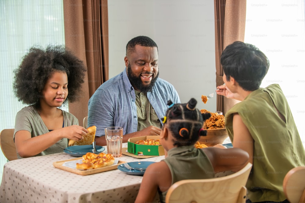 Pais felizes da família africana e duas filhas pequenas comendo frango frito e pizza para o jantar juntos. Pai e mãe e criança fofa menina gostam de comer e compartilhar uma refeição juntos em casa