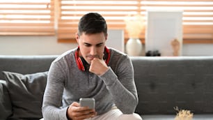 Jeune homme vérifiant les médias sociaux sur son smartphone alors qu’il est assis dans le salon.