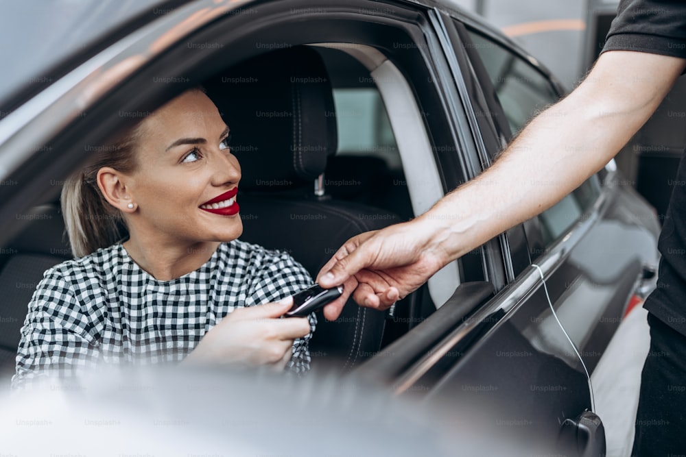 만족스럽고 미소를 지은 여성 구매자는 새 차에 앉아 있습니다. 그녀는 미소를 지으며 열린 창문을 통해 판매자를 바라보며 자동차 열쇠를 가져갑니다.