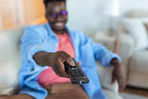 Hombre afroamericano emocionado que apunta el controlador de televisión a la cámara que cambia de canal viendo la televisión. Publicidad de programación televisiva. Enfoque selectivo