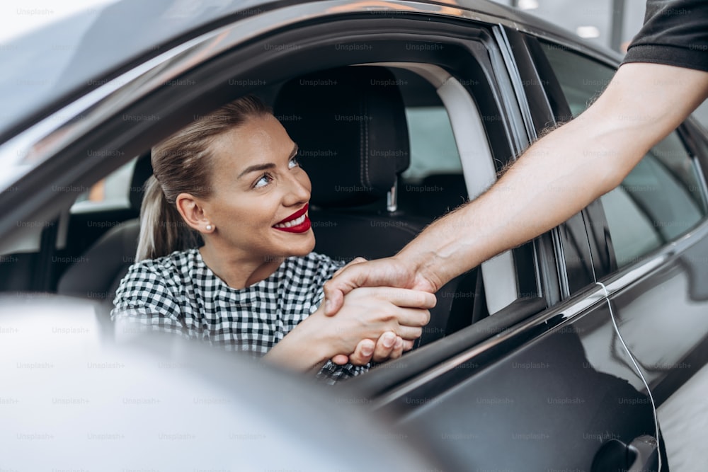 Acquirente femmina soddisfatto e sorridente seduto nella sua nuova auto. Sorride, guarda il venditore attraverso la finestra aperta mentre gli stringe la mano.