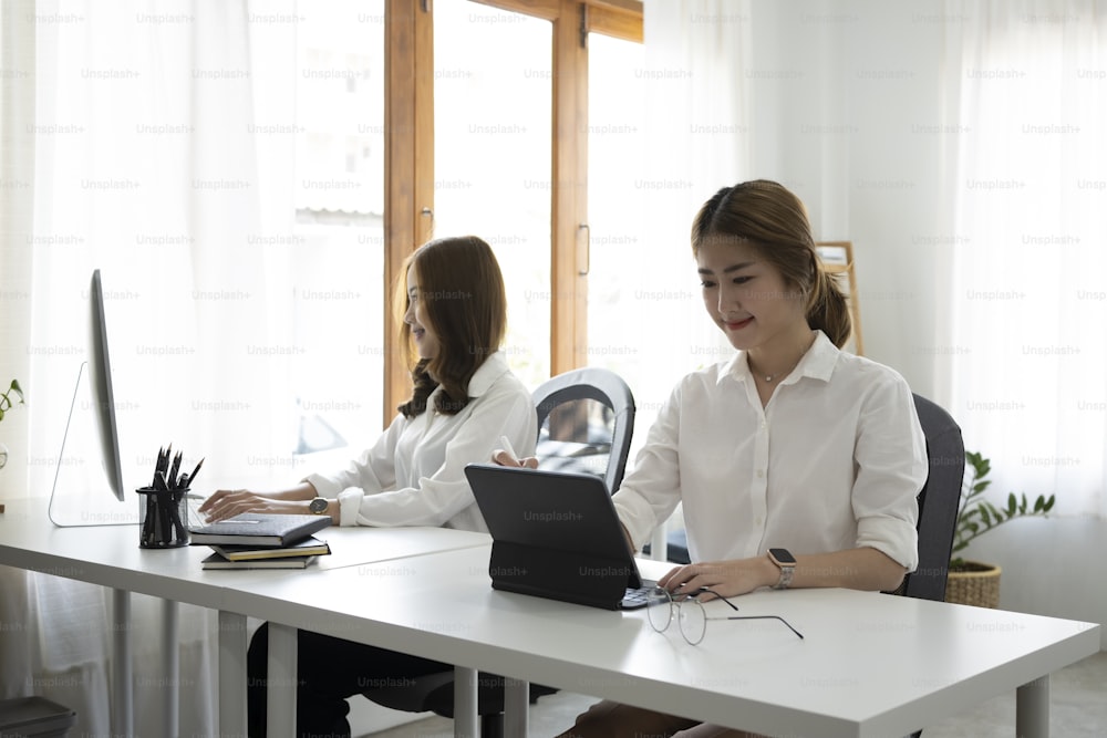 웃고 있는 아시아 여성 직장인은 사무실에서 동료와 함께 앉아 컴퓨터 태블릿으로 작업하고 있다.