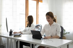Lächelnde asiatische Büroangestellte sitzt mit ihrer Kollegin im Büro und arbeitet mit einem Computer-Tablet.