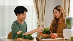 Happy asian couple making fresh green detox vegetable juice in the kitchen.