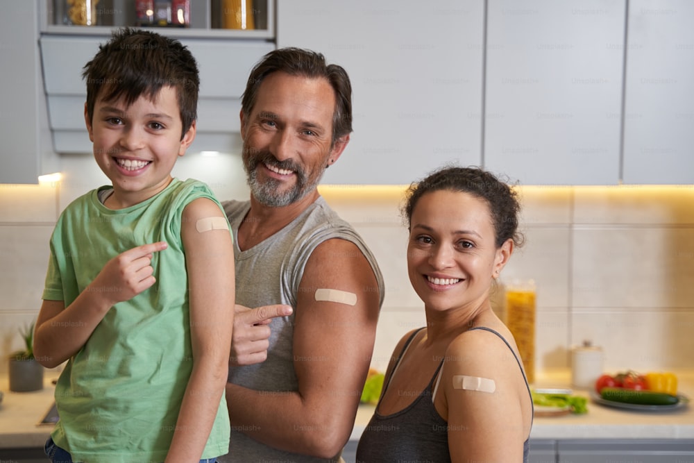 Female with her husband and son looking at camera while demonstrating their adhesive bandages on shoulders in kitchen