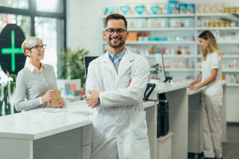 Young female pharmacist posing while working in a pharmacy while his female colleague is selling prescription medications to senior female customer