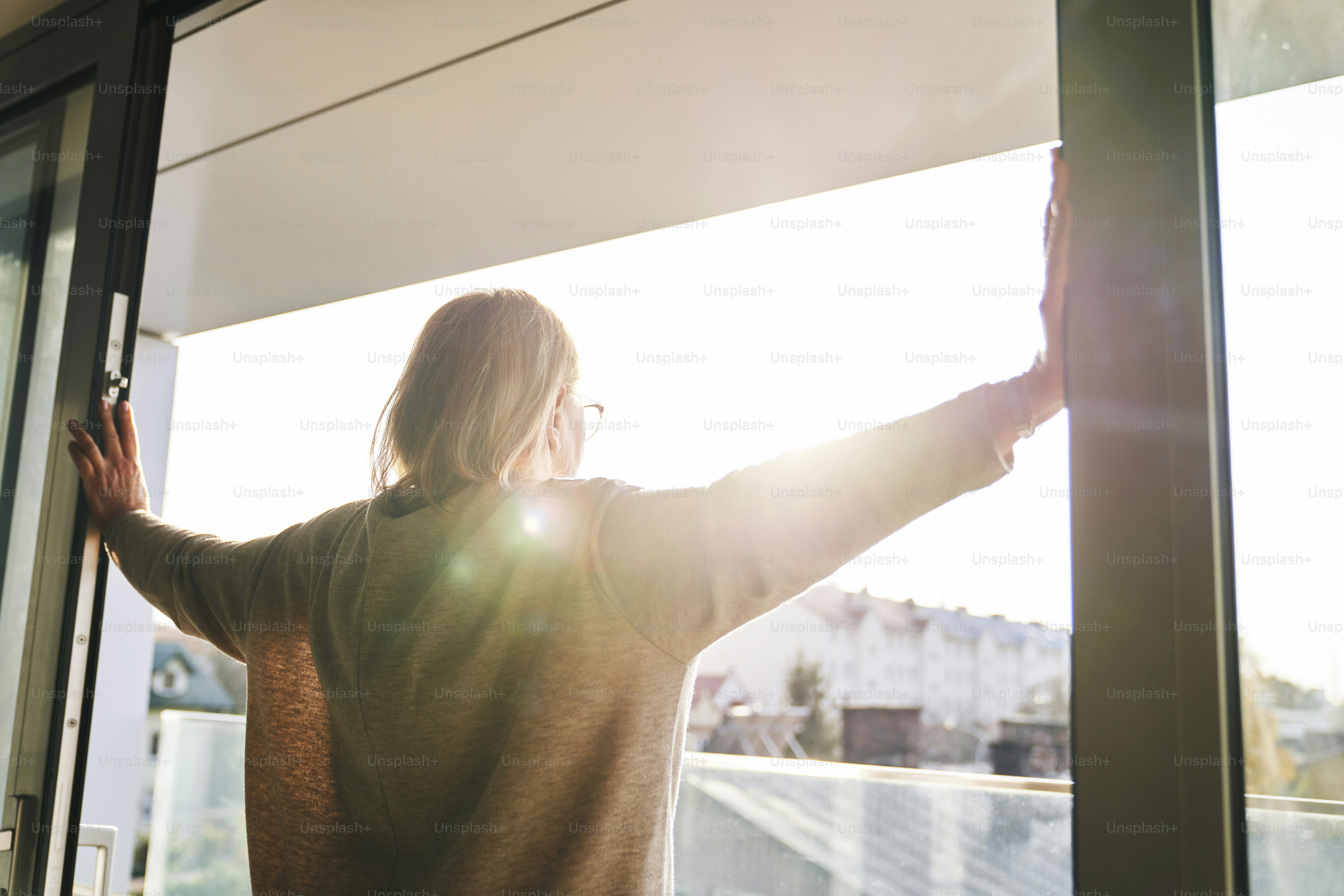Vista trasera de una mujer mayor que abre las puertas del balcón en los rayos del sol