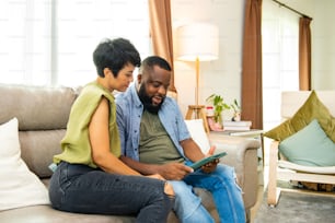 Afrikanisches Paar sitzt auf dem Sofa im Wohnzimmer mit digitalem Tablet für Online-Shopping mit Internet-Banking zusammen. Glückliche Familie entspannen Sie sich und genießen Sie Indoor-Lifestyle-Aktivitäten mit Technologie zu Hause