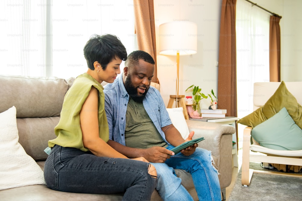 African couple sitting on sofa in living room using digital tablet for online shopping with internet banking together. Happy family relax and enjoy indoor lifestyle activity with technology at home