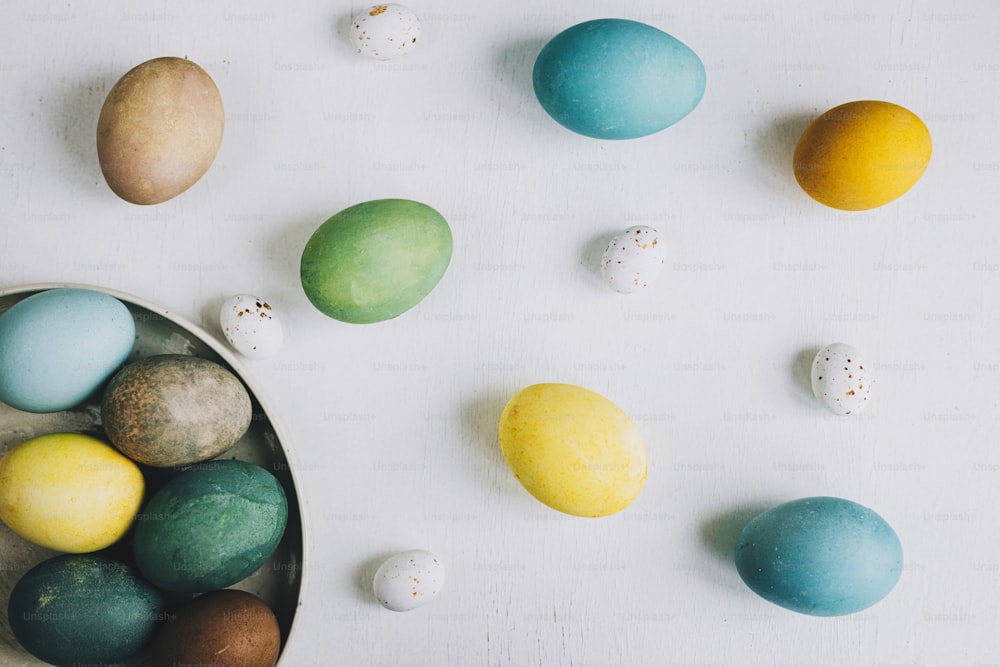 ¡Felices Pascuas! Elegantes huevos de Pascua en plato sobre fondo rústico de madera blanca, planos. Composición de huevos de colores teñidos naturales en mesa rústica