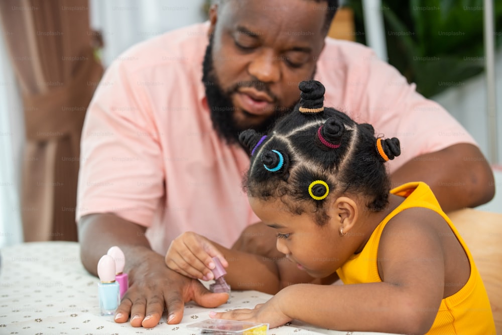 Happy African family father applying nail polish to little daughter in living room. Dad and child girl kid enjoy and having fun leisure activity spending time together with beauty treatment at home