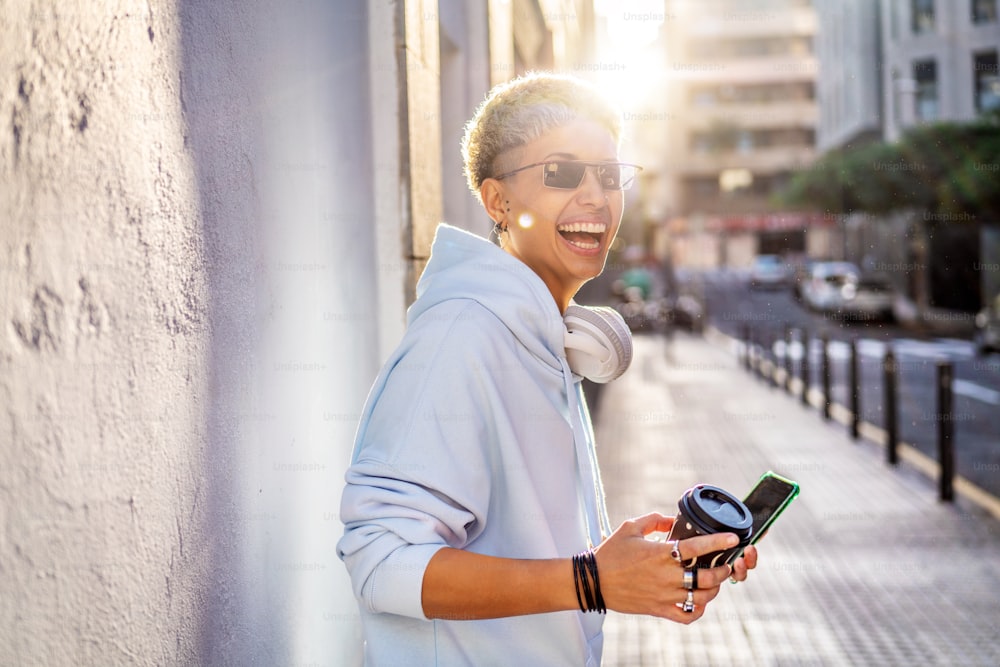 Felice sorridente giovane bella donna ispanica che cammina sulla strada della città con il caffè. Ragazza che indossa una felpa con cappuccio blu alla moda, occhiali da sole e molti anelli. Emozioni di persone vere.