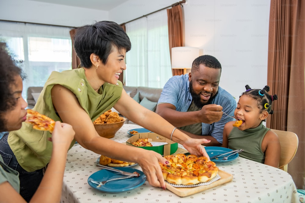 Glückliche afrikanische Familieneltern und zwei kleine Tochter, die zusammen gebratenes Hühnchen und Pizza zum Abendessen essen. Vater und Mutter und süßes Kind genießen es, zu Hause zu essen und eine Mahlzeit zu teilen
