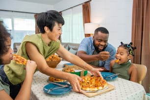Happy African family parents and two little daughter eating fried chicken and pizza for dinner together. Father and mother and cute child girl kid enjoy eating and sharing a meal together at home