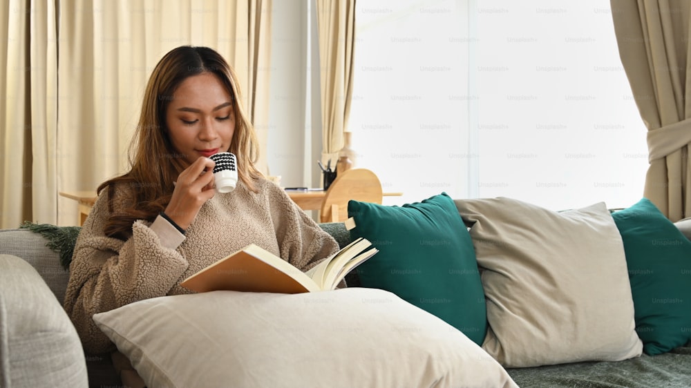 평화로운 아시아 여자가 책을 읽고 소파에서 커피를 마시고 있다.