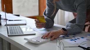 Mujer joven sosteniendo una tarjeta de crédito y usando una calculadora en su lugar de trabajo.