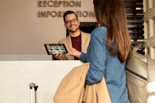 Smiling male receptionist behind the hotel counter showing guest available rooms on tablet.