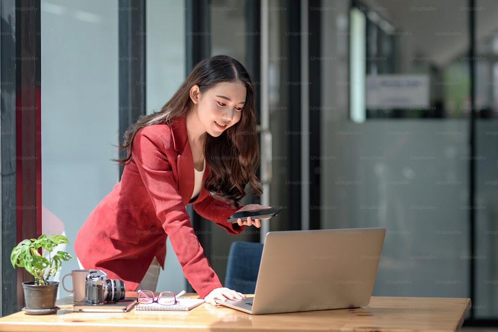 Beautiful young Asian businesswoman standing holding smartphone looking at laptop computer in office.