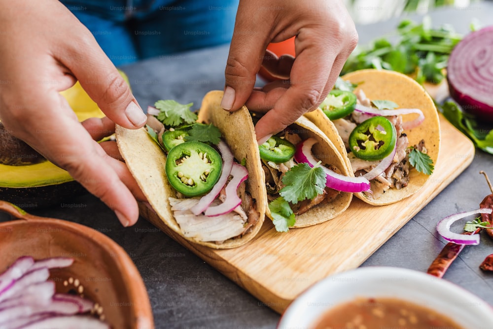 latin woman hands preparing mexican tacos with pork carnitas, avocado, onion, cilantro, and red sauce in Mexico