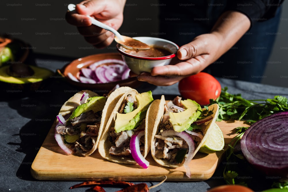 Lateinische Frauenhände bereiten mexikanische Tacos mit Schweinefleisch-Carnitas, Avocado, Zwiebeln, Koriander und roter Sauce in Mexiko zu