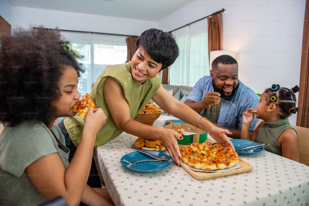 Padres de familia africanos felices y dos hijas pequeñas comiendo pollo frito y pizza para cenar juntos. El padre y la madre y la linda niña disfrutan comiendo y compartiendo una comida juntos en casa