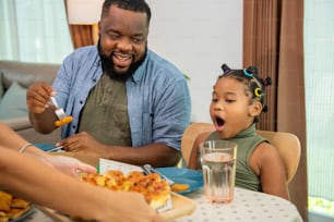 Happy African family parents and two little daughter eating fried chicken and pizza for dinner together. Father and mother and cute child girl kid enjoy eating and sharing a meal together at home