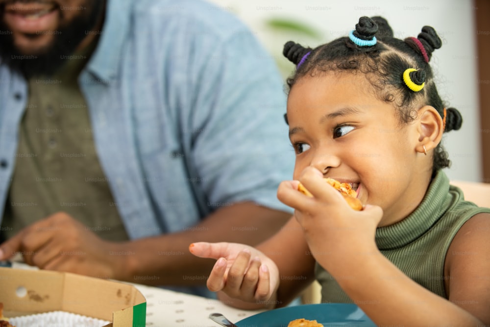 Close up face of African child girl kid biting and eating tasty pizza with happiness. Happy family parents with little cute daughter enjoy having dinner eating and sharing a meal together at home