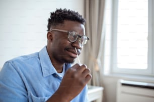 自宅でコロナウイルスPCR検査をしながら綿棒を使用するアフリカ人男性のショット。コロナウイルス迅速診断検査を使用している男性。COVID-19のために鼻腔ぬぐい液を使用している自宅での若い男性。