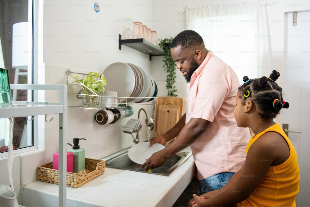African father teaching little daughter washing dishes in the sink on kitchen counter with talking together after having dinner. Happy family dad and child girl kid spending time together at home.
