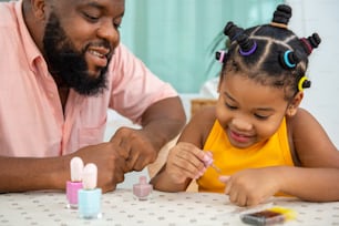 Happy African family father applying nail polish to little daughter in living room. Dad and child girl kid enjoy and having fun leisure activity spending time together with beauty treatment at home