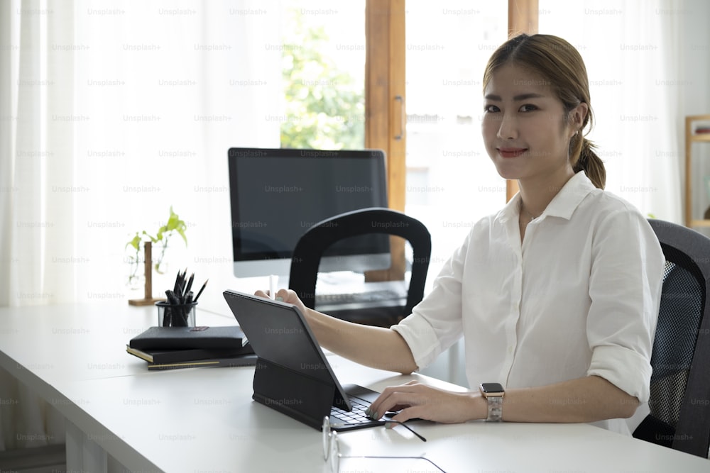 Donna asiatica attraente seduta sul posto di lavoro moderno e sorridente alla telecamera.