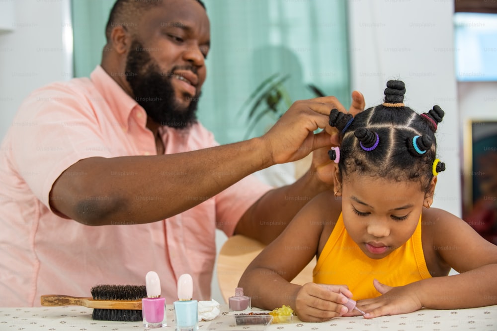 Happy African family father tying little daughter hair with colorful hair band in living room. Dad and child girl kid enjoy and fun spending time together indoor activity with beauty treatment at home