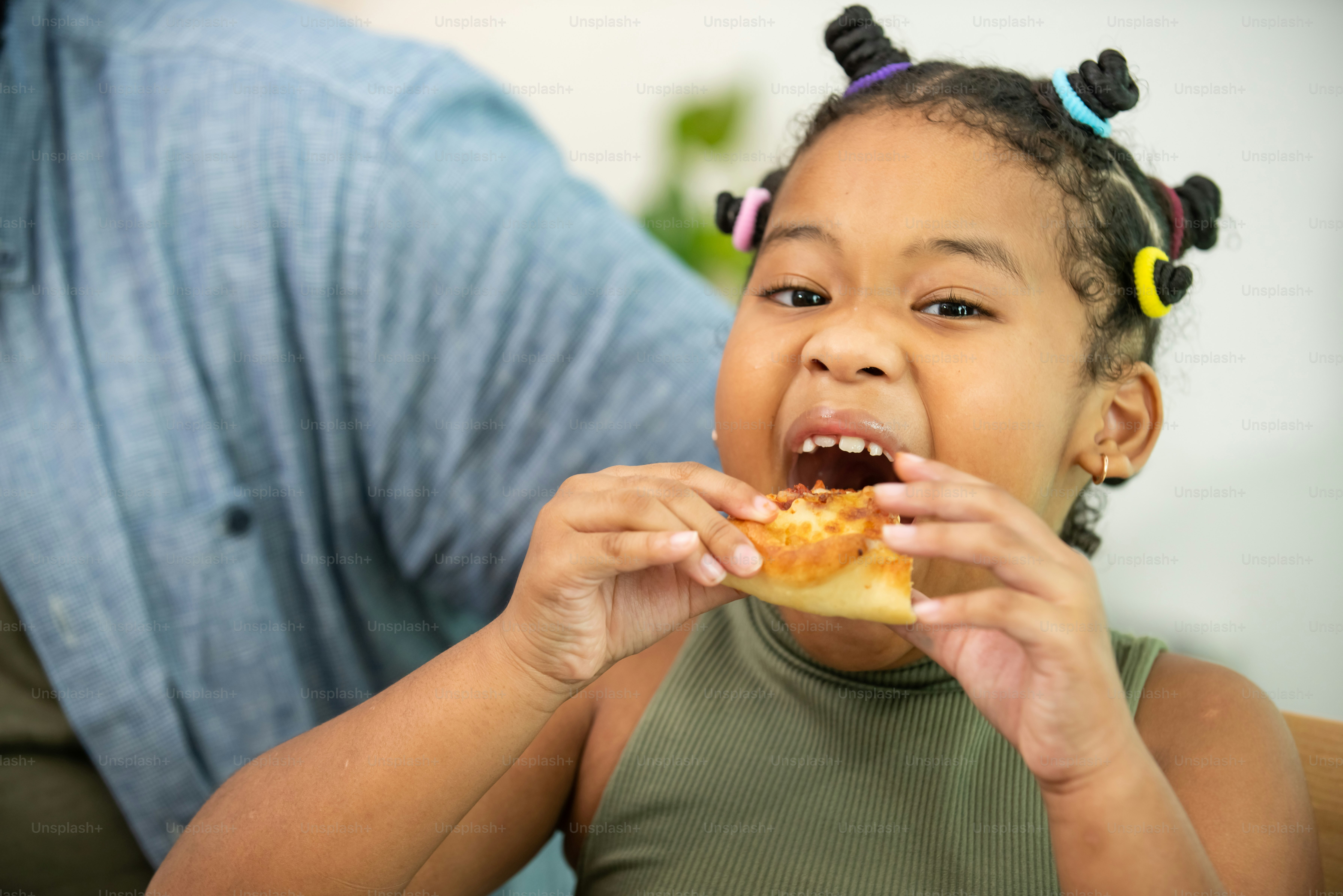 Close up face of African child girl kid biting and eating tasty pizza with happiness. Happy family parents with little cute daughter enjoy having dinner eating and sharing a meal together at home