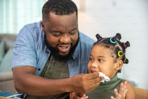 Happy African family parents and two little daughter eating fried chicken and pizza for dinner together. Father and mother and cute child girl kid enjoy eating and sharing a meal together at home