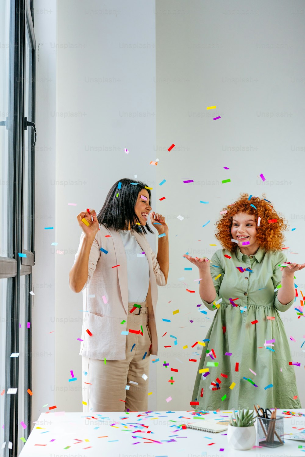 Two young business women having fun time catching confetti in the office. Party time on the work place. Emotions of joy, happiness and delight. Business women celebrating holiday or event in the office. Selective focus.