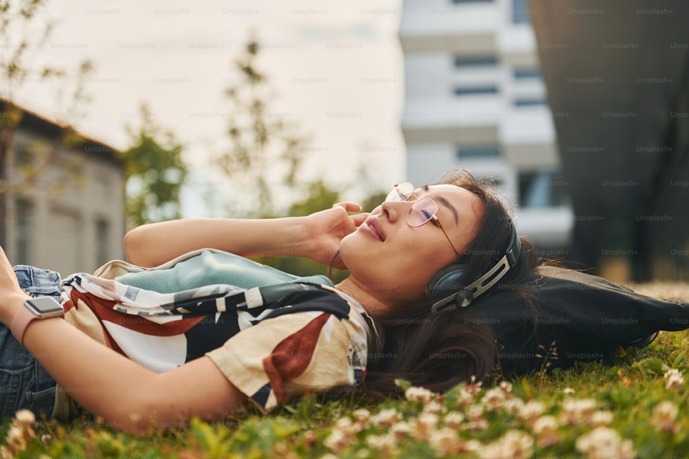 Relaxing on the grass. In headphones. Young asian woman is outdoors at daytime.
